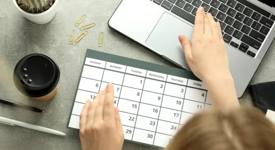 Curso de pós-graduação EaD: qual é a carga horária? | Foto superior de uma mulher mexendo no notebook e olhando um calendário | FAVENI