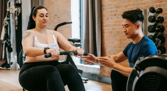 Educação Física na promoção da saúde: qual é a importância? | Foto de personal trainer ajudando aluna a treinar na academia | FAVENI